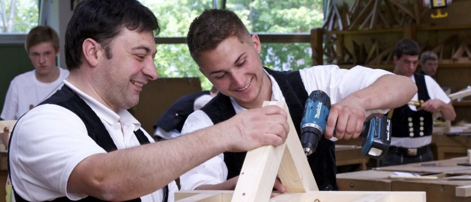 Zimmerer Meister zeigt Schüler wie man zwei Holzteile vertikal miteinander verbindet.