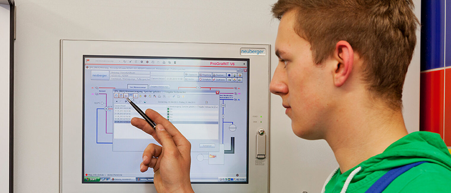 Ein junger Anlagenmechaniker steht vor einer großen Maschine und weist mit einem Stift auf einen darin integrierten Bildschirm.