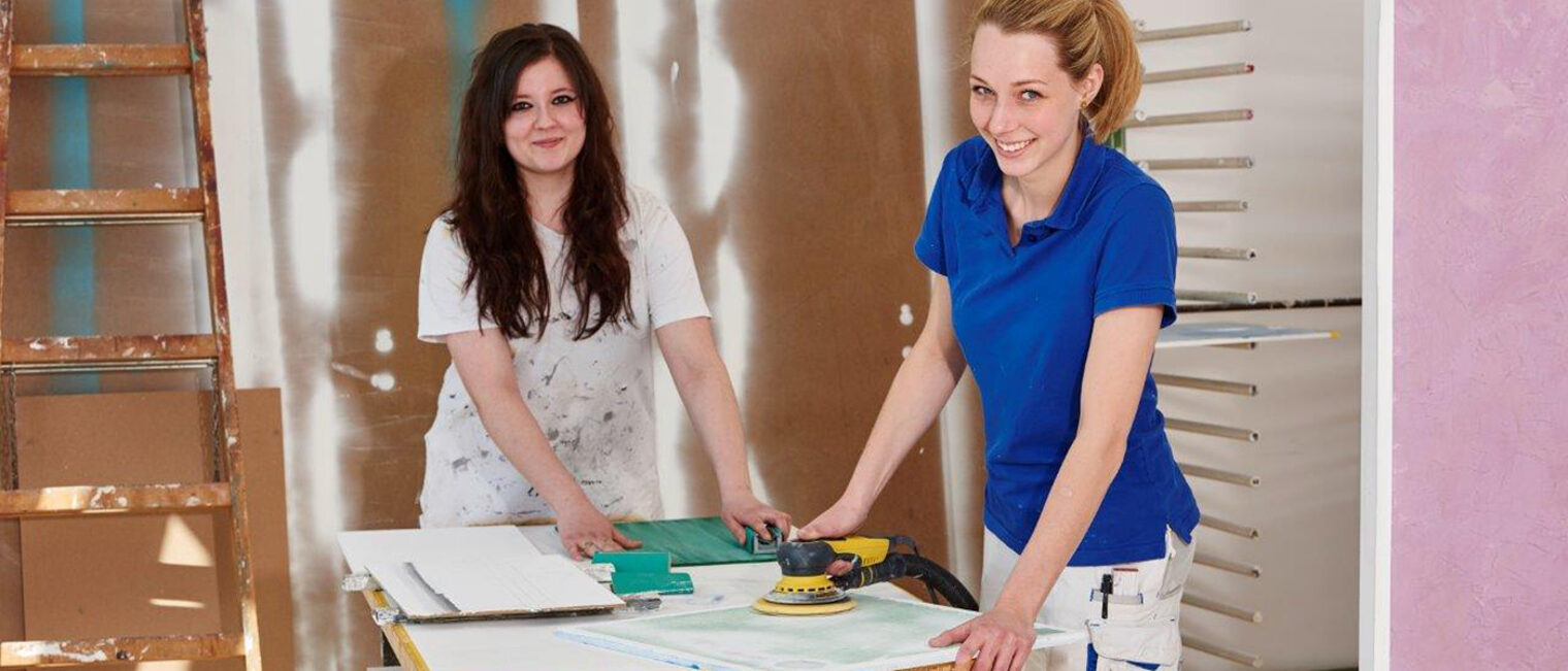 Zwei junge Malerinnen und Lackiererinnen bei der Arbeit lächeln in die Kamera.