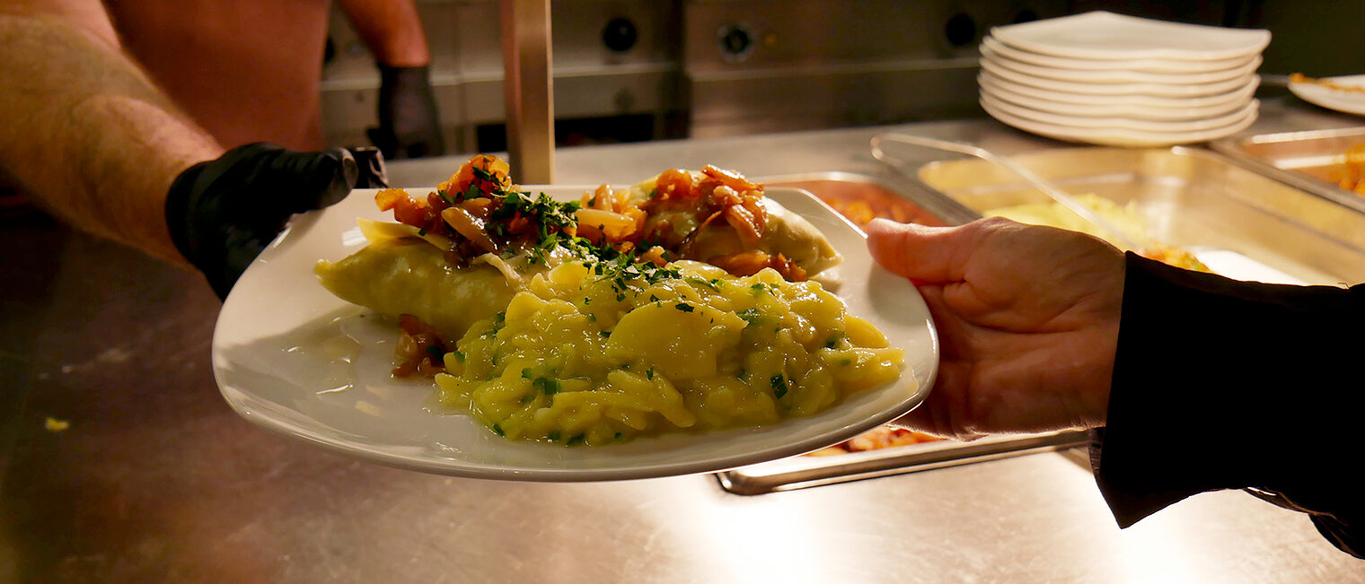 Koch reicht Gast einen Teller mit Maultaschen und Kartoffelsalat