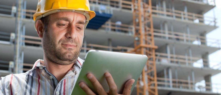 Bauarbeiter mit gelbem Helm schaut in ein Tablet. Im Hintergrund ist eine Baustelle zu sehen.